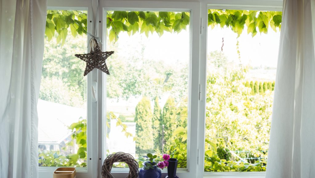Casement windows overlooking a yard covered in trees and bushes. A wooden star and wreath, as vases, adorn the windowsill.