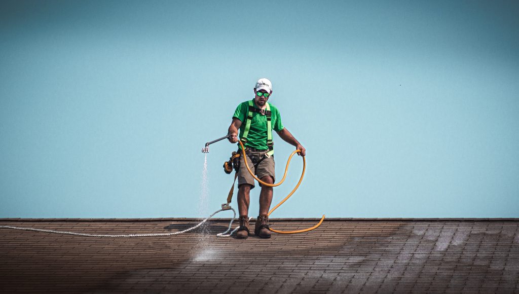A certified Roof Maxx dealer stands on a roof with a hose in hand, applying the Roof Maxx treatment to asphalt shingles.