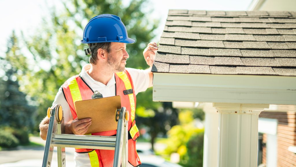 A professional roofer inspecting the edges of a roof. He wears a blue hard hat and high-vis vest, and is standing on a ladder with a clipboard in hand.