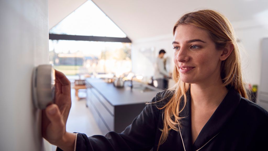 A smiling woman adjusts the smart thermostat in the kitchen of her home.